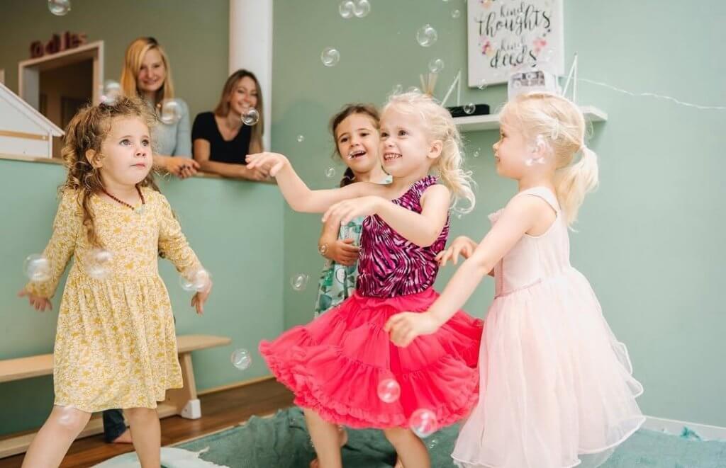 Children dressed as princesses playing in a sensory bubble party, while their mothers are watching and smiling at them.